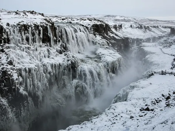 A large waterfall in a snowy canyon, partly frozen, with a viewing path along the edge