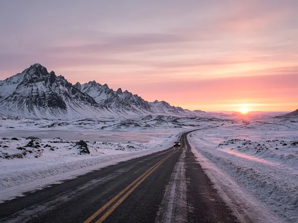 An empty road winding through snowy mountains toward a low pink winter sun