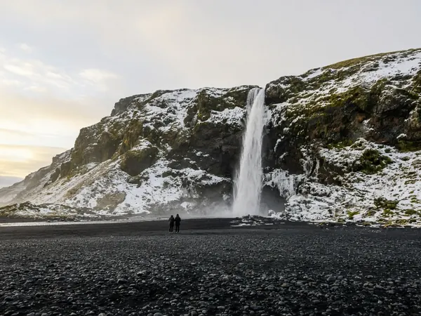 A tall waterfall dropping from a snowy cliff onto a black pebble plain, two people for scale